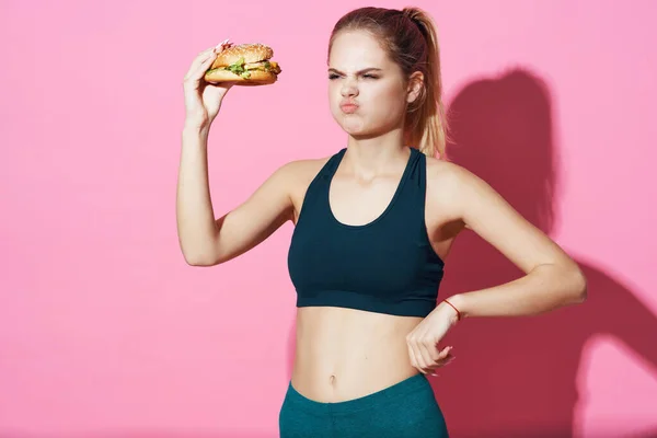 Mujer deportiva con hamburguesa en las manos comiendo comida rosa fondo comida rápida — Foto de Stock