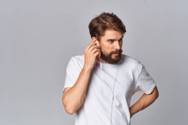 Beyaz tişörtlü, sakallı, günlük giyimli stüdyo ışığı arka planlı bir adam.