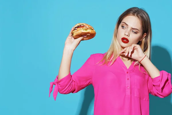Wesoła blondynka w różowej koszuli hamburger fast food snack niebieskie tło — Zdjęcie stockowe