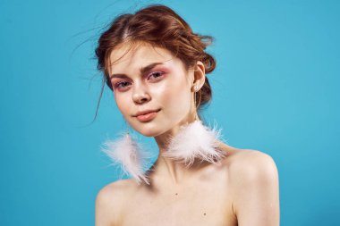 Çıplak omuzlu, parlak makyajlı ve tüylü küpeli bir kadın. Mavi arka plan yakın plan.