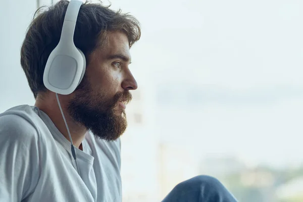 Uomo barbuto seduto vicino alla finestra in cuffia ad ascoltare musica — Foto Stock