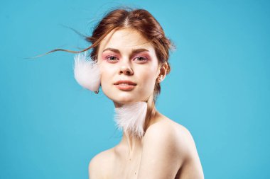 Çıplak omuzlu, parlak makyajlı ve tüylü küpeli bir kadın. Mavi arka plan yakın plan.