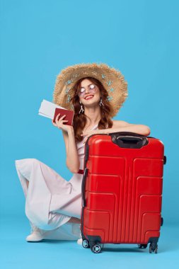Kırmızı valizli kadın pasaport uçak biletlerinin olduğu katta oturuyor mavi arka planda seyahat ediyor.