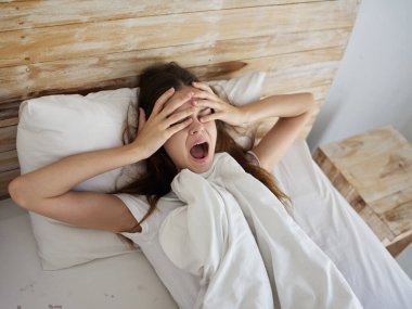 Kadın yatakta ağzı sonuna kadar açık uzanıyor. Sabah ellerini yüzünde tutuyor.