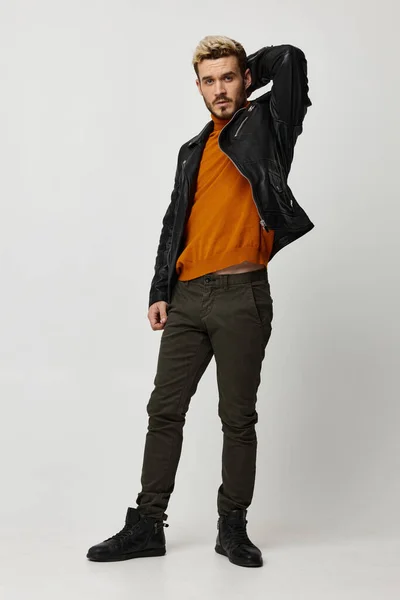Sexy man in a leather jacket holds his hand behind his head and light background orange sweater pants — Stock Photo, Image