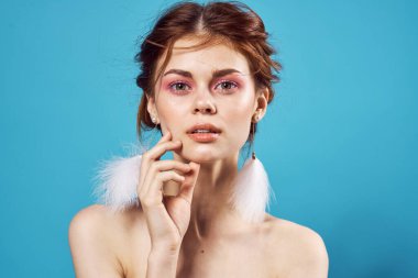 Güzel bir kadın, çıplak omuzlu, tüylü küpeli, saf deri, parlak, mavi arka planlı.