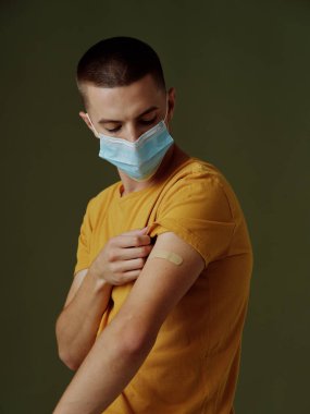 Omzunda yara bandı olan sarı tişörtlü bir adam. 19 numaralı medikal maske güvenlik aşısı.