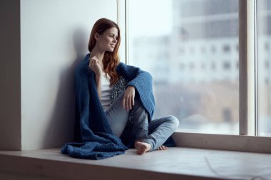 Pencere kenarında oturan mavi ekoseli, rüya gibi bakışlı kadın.