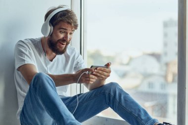 Beyaz tişörtlü adam pencere eşiğinde kulaklıklı ve kulaklıklı oturuyor.
