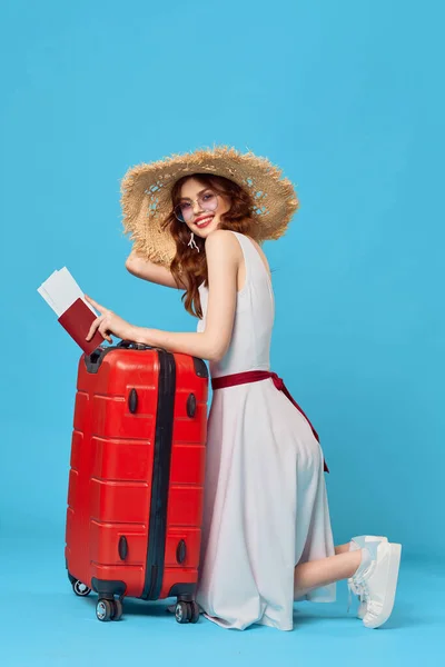Ładna kobieta w kapeluszu z czerwoną walizką siedzi na podłodze niebieskie tło podróży — Zdjęcie stockowe