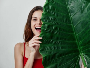 Kırmızı mayo giymiş neşeli bir kadın yüzünün yarısını yeşil bir yaprak tutuyor.