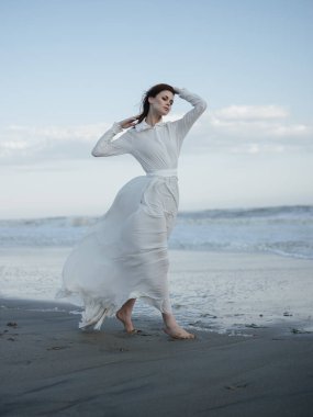 Beyaz elbiseli bir kadın kumların üzerinde yalın ayak yürüyor.