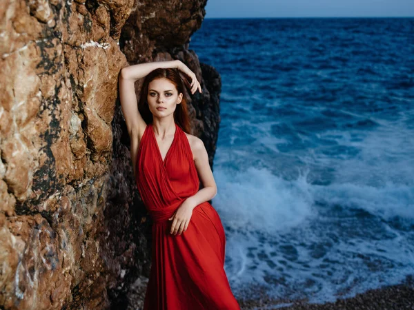 Ελκυστική γυναίκα σε κόκκινο φόρεμα από τους βράχους του ωκεανού ποζάρουν — Φωτογραφία Αρχείου