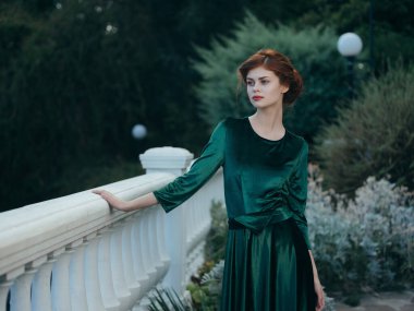 Güzel bir kadın yeşil bir elbise parkında korkuluklara yaslanıyor.