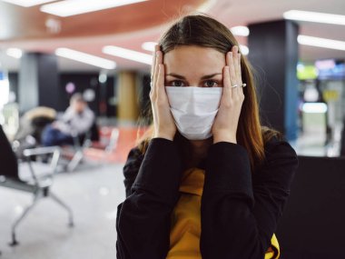 Havaalanında oturan kadın tıbbi maske takıyor ve uçuş beklerken yüzünü tutuyor.