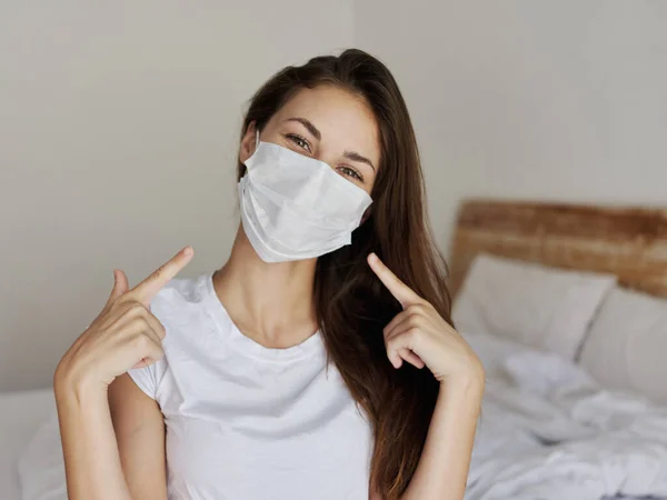 Счастливая женщина, сидящая на кровати и показывающая пальцем на медицинскую маску на лице — стоковое фото