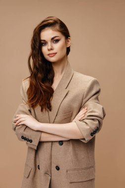 Paltolu şık bir kadın bej bir arka planda kollarını göğsünde kavuşturdu.
