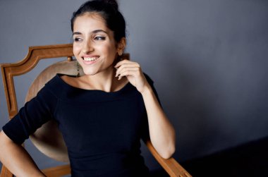 Esmer küpeler sandalyenin yanında poz veren siyah elbiseler koyu arkaplan