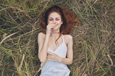 Beyaz elbiseli neşeli kızıl saçlı kadın çimenlerin üzerinde uzanıp dinleniyor.