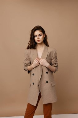 Bej arka planda poz veren şık bir kadın. Palto ve pantolonlu.
