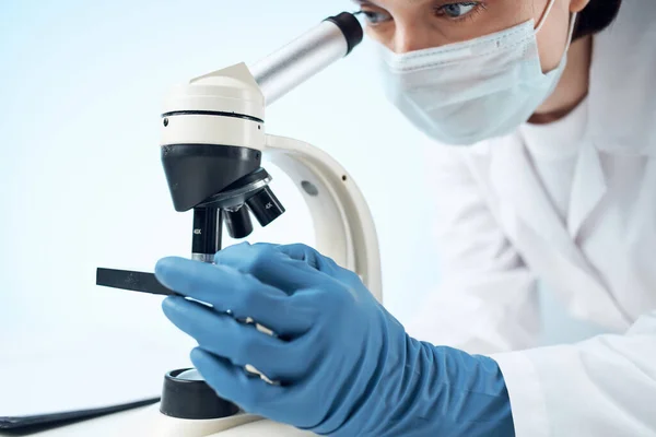 Βοηθός εργαστηρίου φορώντας μια ιατρική μάσκα κοιτάζοντας μέσα από ένα μικροσκόπιο — Φωτογραφία Αρχείου