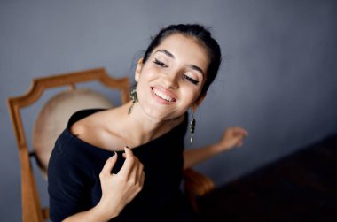 Esmer küpeler sandalyenin yanında poz veren siyah elbiseler koyu arkaplan