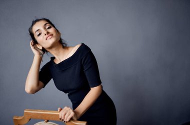 Esmer küpeler sandalyenin yanında poz veren siyah elbiseli yaşam tarzı stüdyosu.