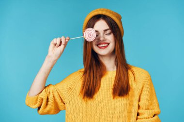 Sarı kazaklı neşeli bir kadın elinde lolipopuyla stüdyoda poz veriyor.