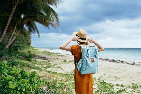 Woman in a sundress and hat on the island near the trees and the sea Backpack in the background — Stock Photo, Image