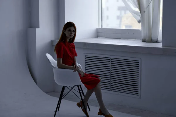 빨간 드레스를 입은 빨간 머리 여자가 창문 옆 의자에 앉아 있습니다 — 스톡 사진