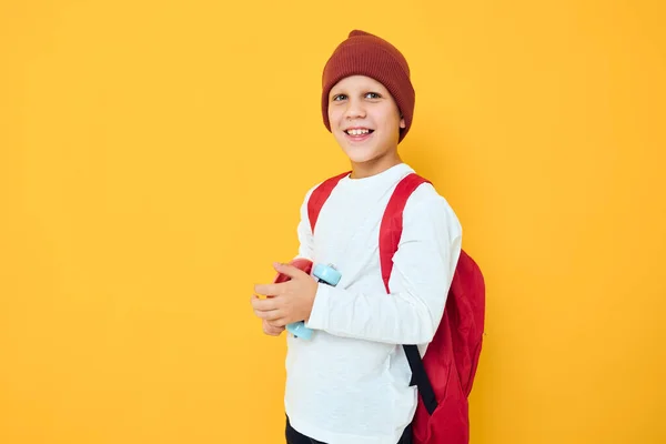 Criança alegre com mochila vermelha skate vermelho cor amarela fundo — Fotografia de Stock