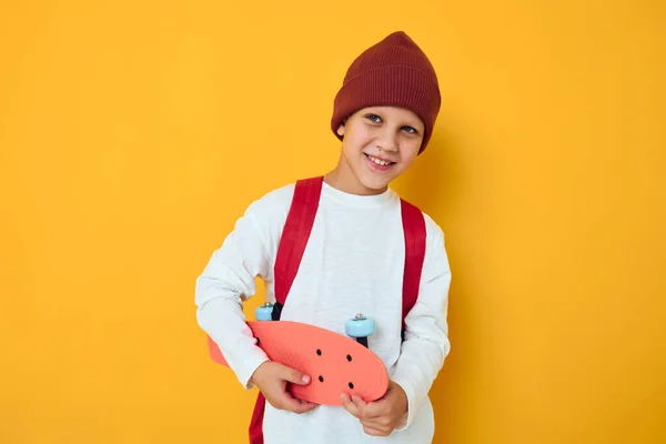 Glad skolpojke i en röd hatt skateboard i händerna gul färg bakgrund — Stockfoto