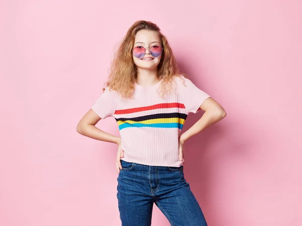 안경을 쓰고 티셔츠를 입고 핑크 색 배경을 가진 멋진 소녀가 허리에 손을 잡고 있습니다. — 스톡 사진