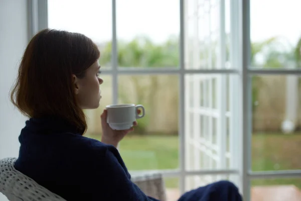 Kobieta siedząca w domu i wyglądająca przez okno filiżankę kawy — Zdjęcie stockowe