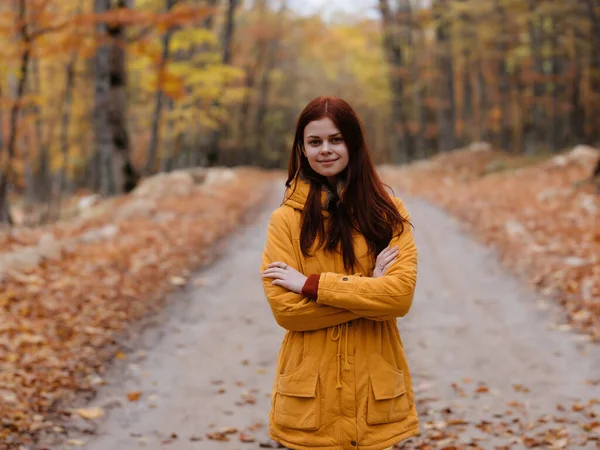 Sarı ceketli bir kadın sonbahar orman yolunda yürüyor.