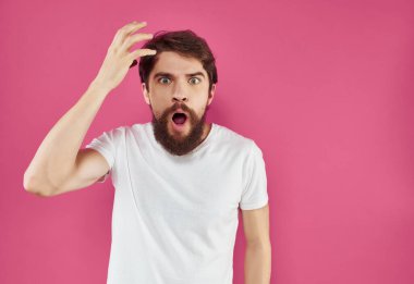 Beyaz tişörtlü sakallı adam pembe arka planda el hareketi yapıyor.