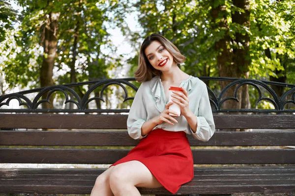 Mujer alegre sentada en un banco con una taza de café al aire libre en el parque — Foto de Stock