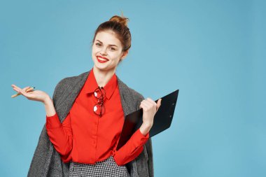 İş kadını, omuzlarında ceket, elinde klasörle mavi arka planda çalışıyor.