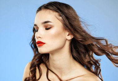 Çıplak omuzlu, kırmızı dudaklı, temiz tenli, mavi arkaplan pozu veren bir kadın.