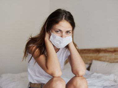 Kızıl saçlı, tıbbi maskeli bir kadın tecrit odasında yatağında oturuyor.