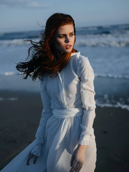 Όμορφη γυναίκα σε λευκό φόρεμα που θέτουν πολυτέλεια με τα πόδια κατά μήκος της παραλίας — Φωτογραφία Αρχείου