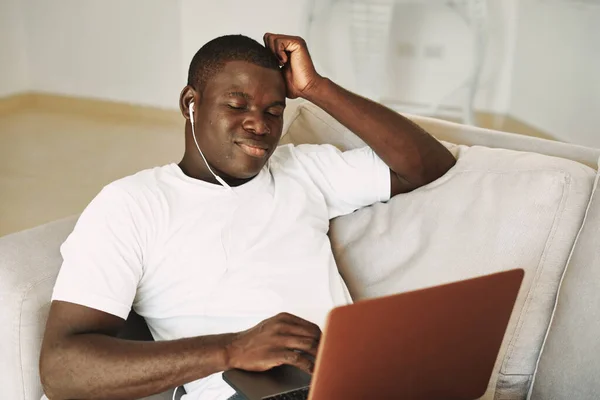 a man of african appearance with a closed laptop sits on the couch and listens to music on headphones