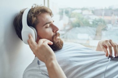 Beyaz tişörtlü adam pencere eşiğine uzanmış kulaklık takıyor.