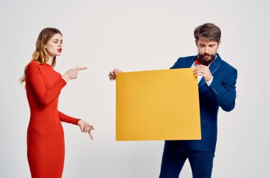 Takım elbiseli bir iş adamı kırmızı elbiseli bir kadının yanında sarı model posterler asıyor.