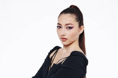 Keskin pembe göz makyajı ve uzun esmer at kuyruklu, beyaz arka plana karşı şık giyinen çekici bir model. Makyajda ve modada yaratıcılığı vurguluyor.