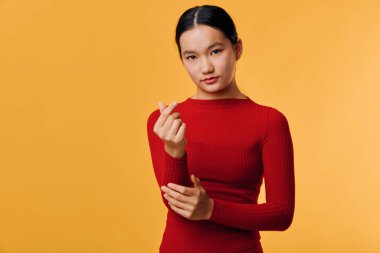 Kırmızı uzun kollu bluz giyen genç Asyalı kadın turuncu arka planda parmak kalp hareketi gösteriyor. Stüdyoda sakin bir ifadeyle ve doğrudan göz teması kurarak poz veriyor..