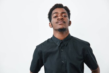 Siyah gömlekli, siyah tenli, beyaz arka planda poz veren kendinden emin bir Afrikalı. Erkek portresi, doğal saçlar ve kameraya bakarak sakince yüz ifadesi..