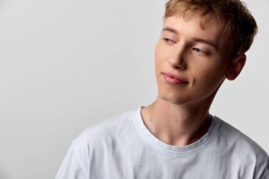 Beyaz tişörtlü genç bir adam yan tarafa bakarken rahat bir tarz. Stüdyo ışığı pürüzsüz cilt tonlarını artırıyor. Günlük kullanım için en az kompozisyon.