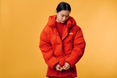 Kırmızı kabarık ceketli genç bir Asyalı kadın canlı sarı stüdyo arkaplanında sıcak bir dış giysi ilikliyor. Mevsimlik kıyafet, portre, günlük kıyafet ve kış modası ayrıntıları.