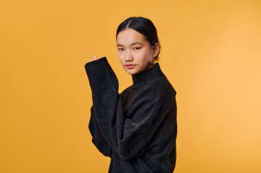 Siyah örülmüş süveter giyen, kolları turuncu arka planda çapraz duran, modaya uygun moda portresi için az makyajlı ve parlak saçlı bir kadın model..